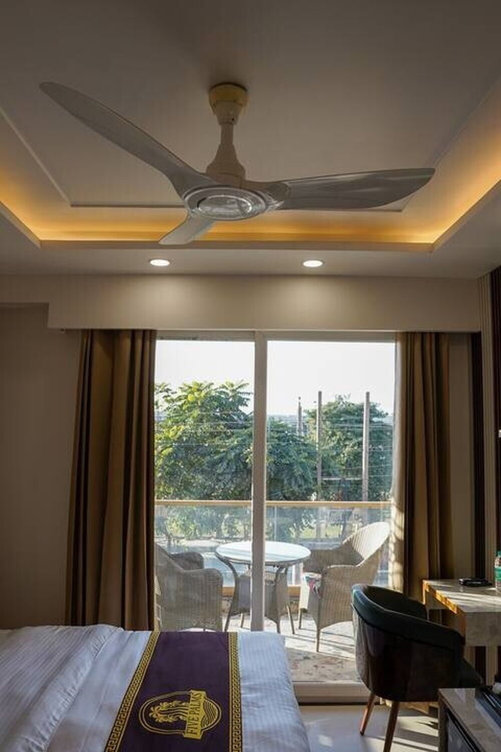 Habitación de hotel con cama, ventilador de techo, balcón con sillas y mesa, y vista a zonas verdes.
