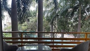 A balcony with wicker furniture, a glass table, and a view of palm trees and buildings.