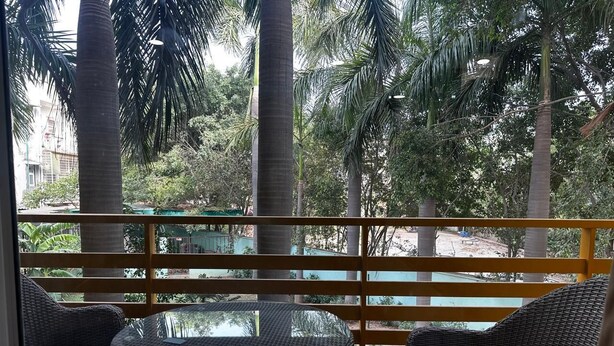 A balcony with wicker furniture, a glass table, and a view of palm trees and buildings. - Five Palms Hotels (Gurugram)