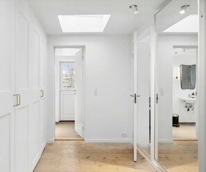 Ein Flur mit weißen Wänden, Holzboden und einem Oberlicht. Mehrere Türen führen in andere Räume, und an der rechten Wand hängt ein Spiegel.