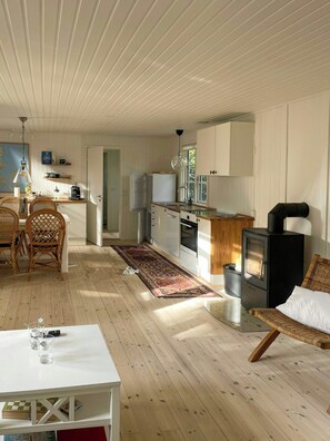A spacious living area with a dining table, wicker chairs, a kitchen with a stove, and a large window. - Summer house with sauna near Udsholt Beach (Græsted)