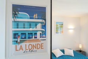 Ein Poster von 'La Londe' zeigt ein Gebäude und Palmen, ein Bett mit weißen Kissen sowie einen Nachttisch mit einer Lampe.
