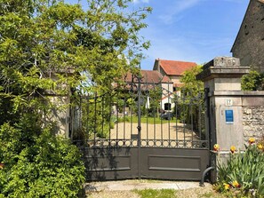 Ein begrenzter Eingang mit einer steinernen Säule, auf der '12' steht, ein Gebäude mit rotem Ziegeldach und ein Garten mit gelben Blumen.
