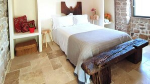 A bedroom with a bed, a bench, a bedside table, a hanging light fixture, and stone walls. - Charming renovated workshop in Ardèche (Joyeuse)