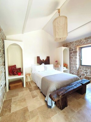 Ein Schlafzimmer mit einem Bett, einer Bank, einem Nachttisch, einer Pendelleuchte und Steinwänden.