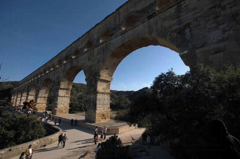 A stone bridge with multiple arches, surrounded by greenery and people walking.