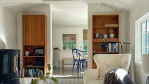 Een ruime woonkamer met een hoge plafon, houten boekenkasten, een comfortabele fauteuil, een eethoek met blauwe stoelen en een keuken die zichtbaar is door een open deur.