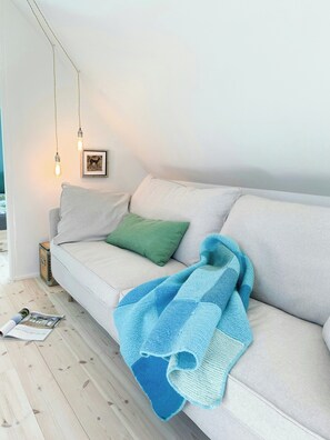 A modern living room with a light-colored sofa, a green pillow, a blue knitted blanket, and two hanging light fixtures.