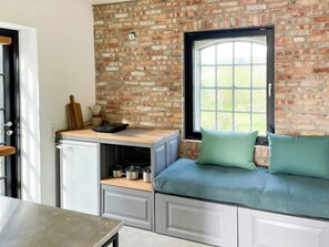 A compact kitchen with a brick wall, a window, a built-in bench with cushions, and a small refrigerator.