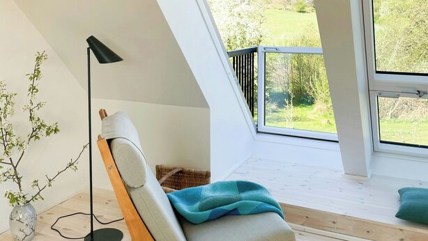 Une chambre moderne avec un plafond en pente, une seule fenêtre donnant sur la verdure, une chaise en bois avec un coussin et un lampadaire.