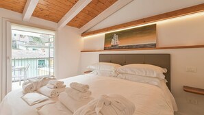 Quarto de hotel com cama, toalhas, pintura de um veleiro e varanda com uma cadeira.