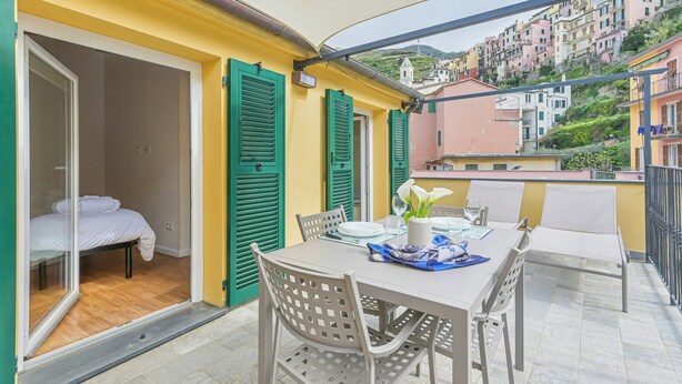 Varanda com mesa posta para refeições, cama no quarto adjacente e vista para edifícios coloridos ao fundo.
