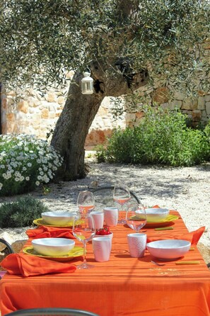 Um espaço de refeições ao ar livre com uma mesa posta para dois, sob uma oliveira, com uma parede de pedra e vegetação ao fundo.