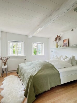 Een slaapkamer met een bed, twee ramen, een radiator, een klein nachtkastje en een zacht, pluizig vloerkleed.
