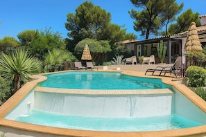 Ein Pool mit gestufter Kante, umgeben von Liegestühlen, einem Reetdachschirm und üppiger Bepflanzung.