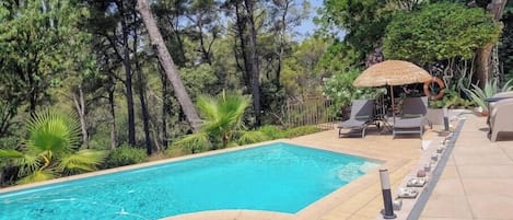 A swimming pool with lounge chairs and a thatched umbrella, surrounded by trees.