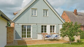 Une maison avec un toit à pignon, de grandes fenêtres et une terrasse aménagée avec du mobilier d’extérieur.