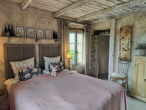 A bedroom with a bed, two pillows, a nightstand, a lamp, a window with curtains, a door, and a wall with framed pictures. - Provençal apartment near Saint-Paul de Vence (Roquefort-les-Pins)
