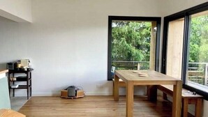 Ein geräumiges Zimmer mit Holzboden, einem großen Fenster mit Blick auf Grünflächen und einem Tisch mit Stühlen.