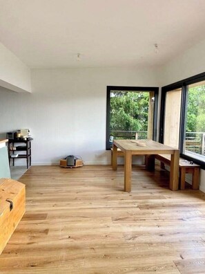 Une pièce spacieuse avec un plancher en bois, une grande fenêtre donnant sur un paysage verdoyant et une table avec des chaises.