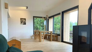 Ein modernes Wohnzimmer mit einem grünen Sofa, einem Holzesstisch und Stühlen. Ein großes Fenster bietet Blick auf viel Grün draußen.