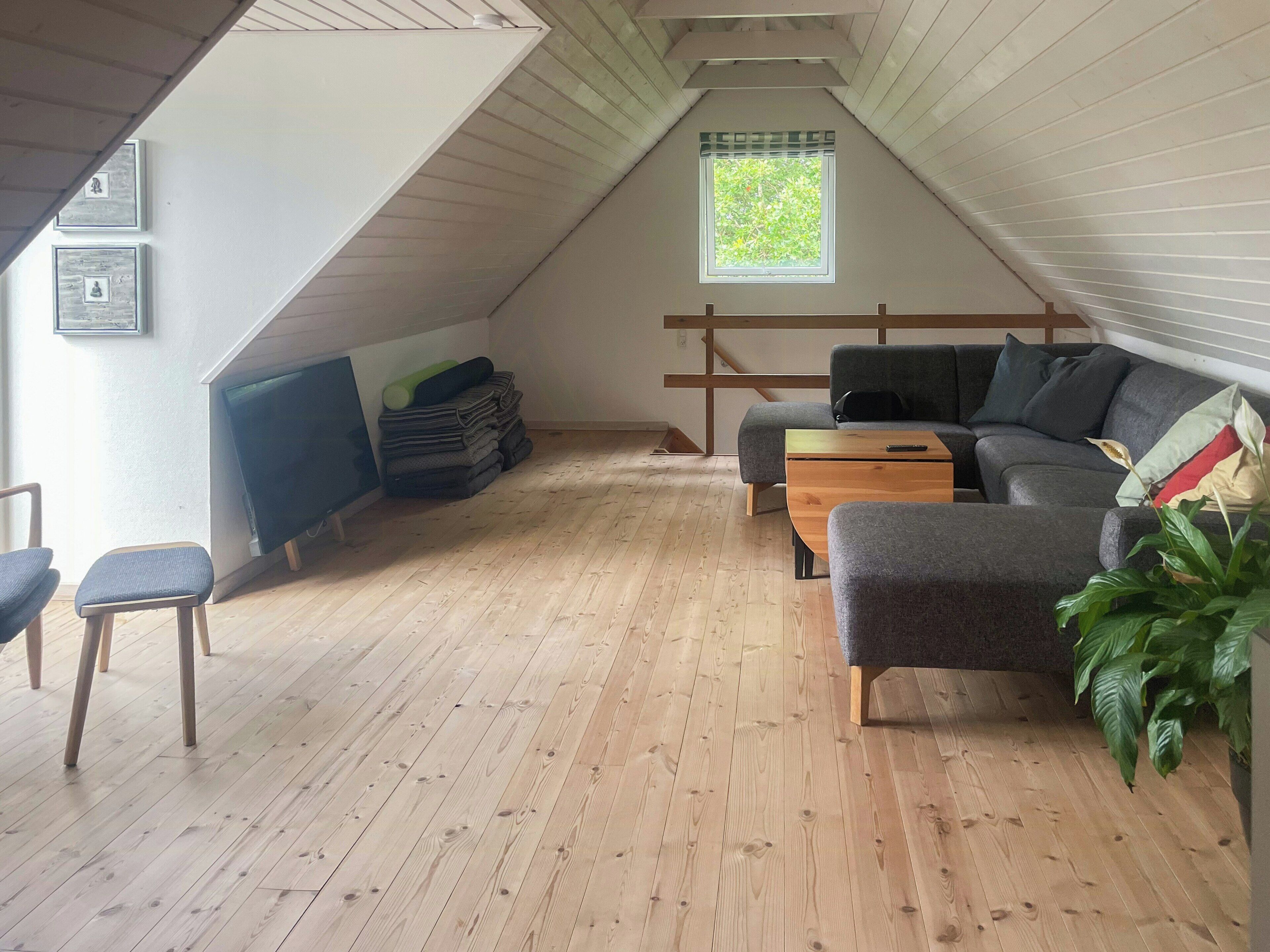 A loft-style room with a sloped ceiling, a wooden floor, a grey sofa, a wooden coffee table, a TV, and a window with a view of greenery.