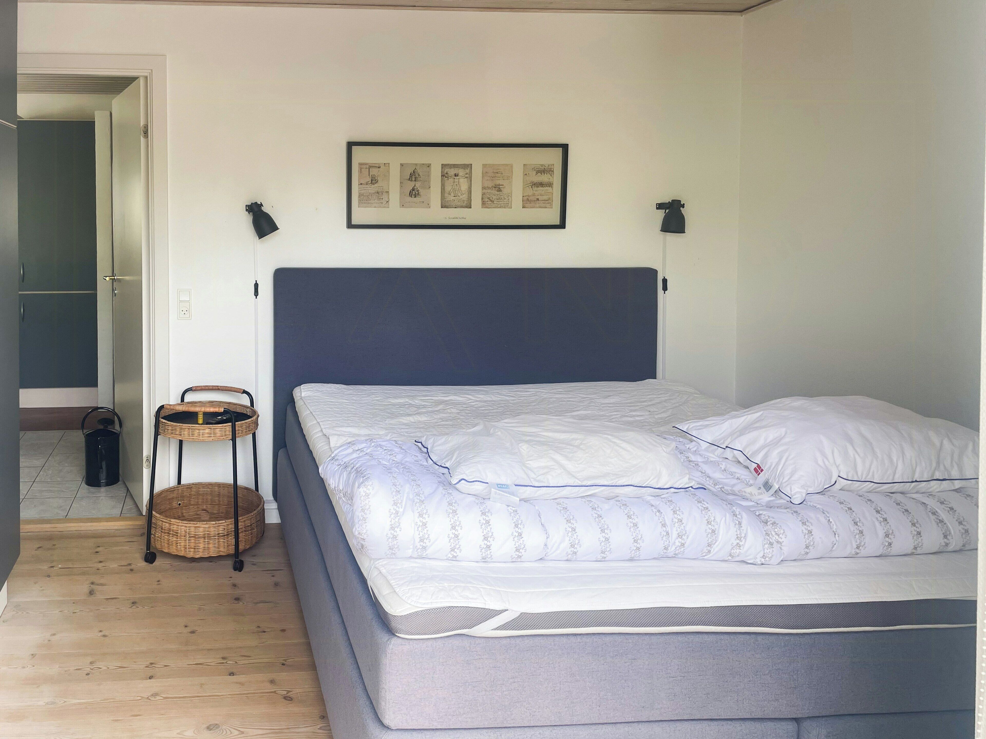 A bedroom with a bed, a framed picture on the wall, a small table, and a basket.