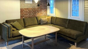 Sala de estar com sofá verde, mesa de centro de madeira, parede de tijolos e duas janelas.