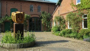 Un patio con un edificio de ladrillo, una escultura de madera y un jardín con diversas plantas.