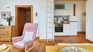 Una sala moderna con un sillón rosa, suelo de madera, un mueble de madera y una cocina con armarios y electrodomésticos blancos.