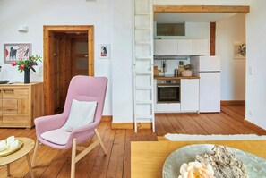 Una sala moderna con un sillón rosa, suelo de madera, un mueble de madera y una cocina con armarios y electrodomésticos blancos.