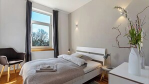 Un dormitorio moderno con cama, una silla, un escritorio, un florero con flores y una ventana con cortinas.