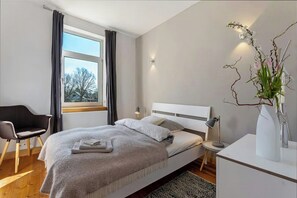 Un dormitorio moderno con cama, una silla, un escritorio, un florero con flores y una ventana con cortinas.