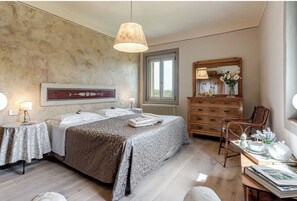 Ein Schlafzimmer mit einem Bett, Nachttischen, einem Schminktisch, einem Sessel und einem Fenster.