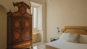 Quarto com cômoda de madeira, cama com roupa de cama branca, radiador e janela.