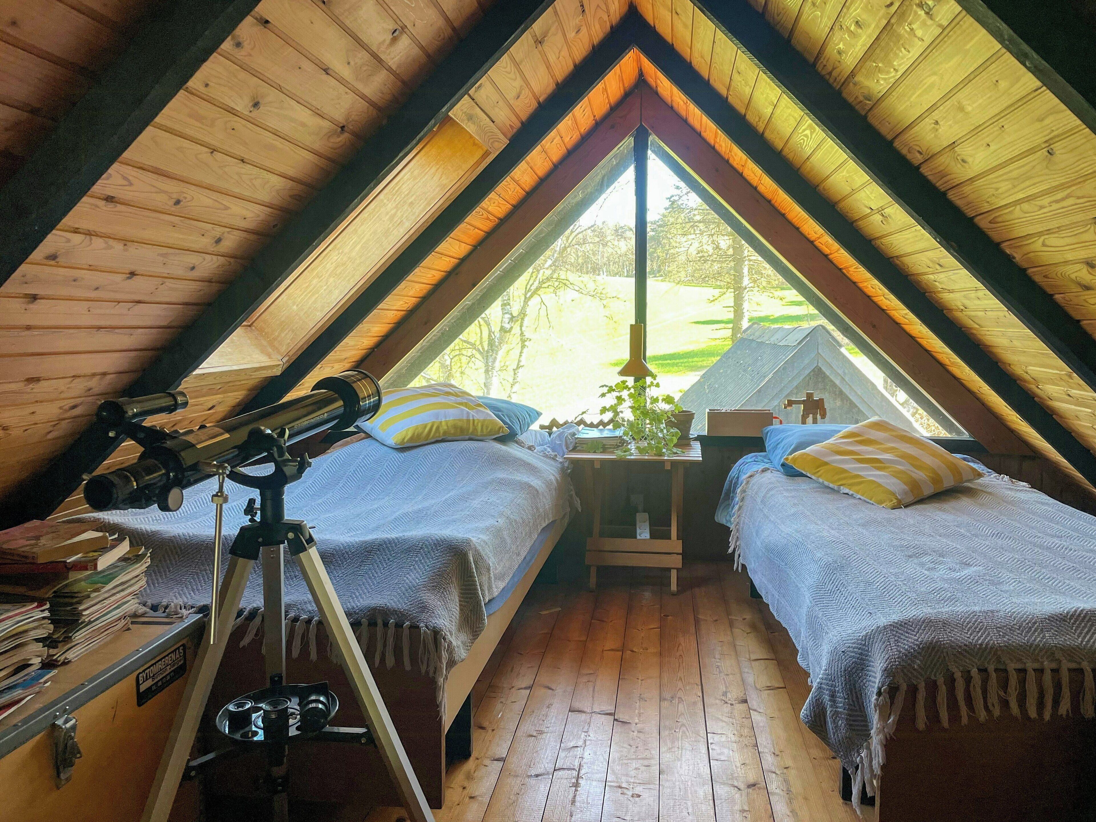 A cozy attic bedroom with two beds, a telescope, and a view of the outdoors.