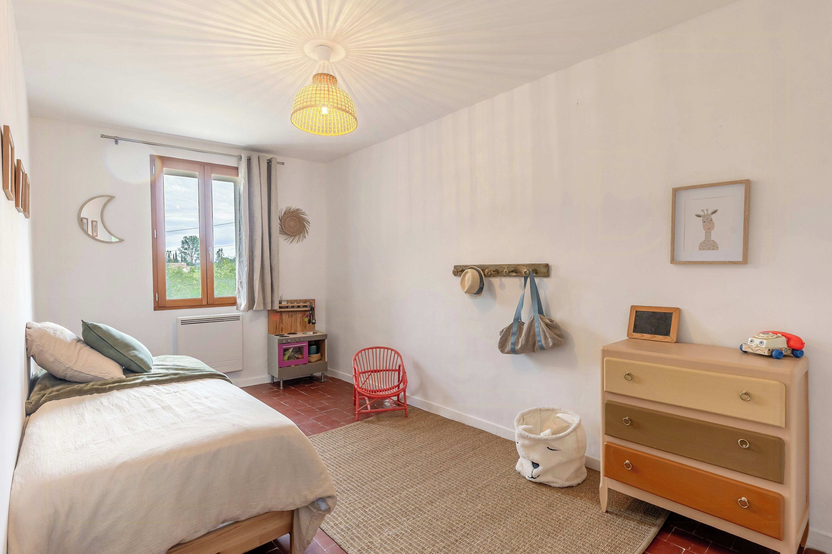 Ein Schlafzimmer mit einem Bett, einem Schminktisch, einem Sessel, einer Pendelleuchte und einem Fenster mit Vorhängen.