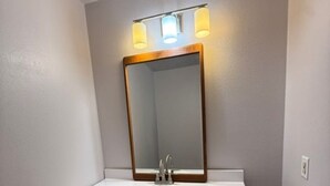 A bathroom with a white vanity, a mirror with a wooden frame, and a wall-mounted light fixture. - The Village Motel (Cambridge)