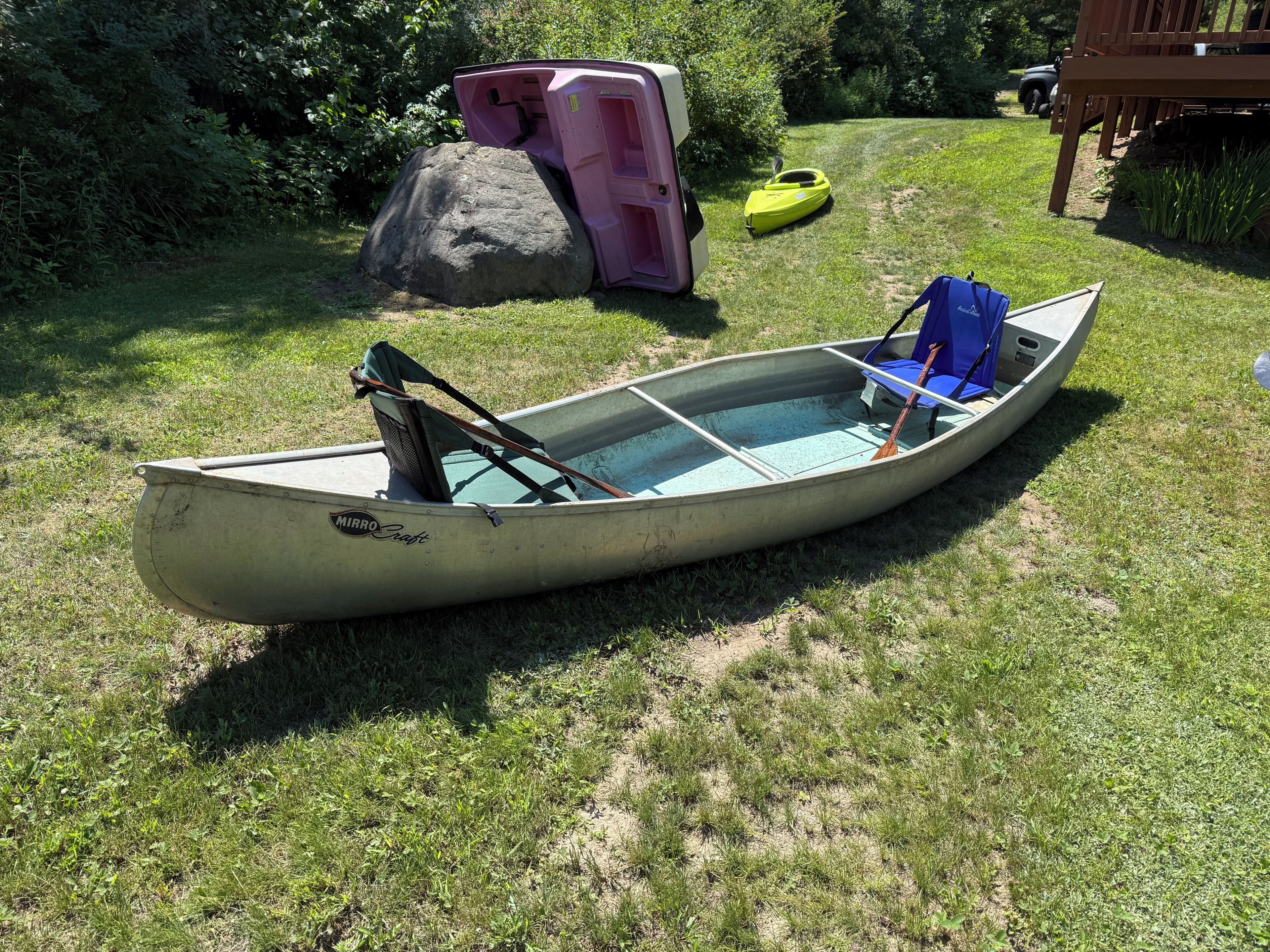 Ein Ruderboot mit blauer Sitzbank und Ruder, ein Kajak und ein pinkes Spielhaus auf einer grünen Wiese.