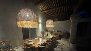 Uma sala de jantar com uma longa mesa de madeira, luminárias pendentes de vime e uma cozinha ao fundo.