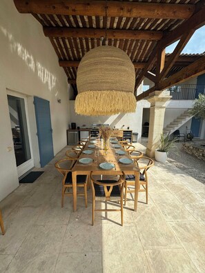 Ein Essbereich im Freien mit einem Holztisch, gedeckt für sechs Personen, umgeben von Rattan-Stühlen unter einer reetgedeckten Überdachung.