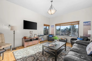A living room with a flat-screen TV, a sofa, a coffee table, a floor lamp, and a large window with a view of trees.