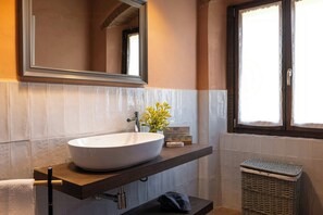 Ein Badezimmer mit einem großen weißen Waschbecken, einem Spiegel darüber, einem Fenster mit Vorhang und einem Korbwannen-Korb.