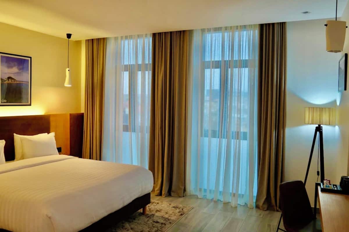 A hotel room with a large bed, a desk, a lamp, and a view of the outdoors through sheer curtains.