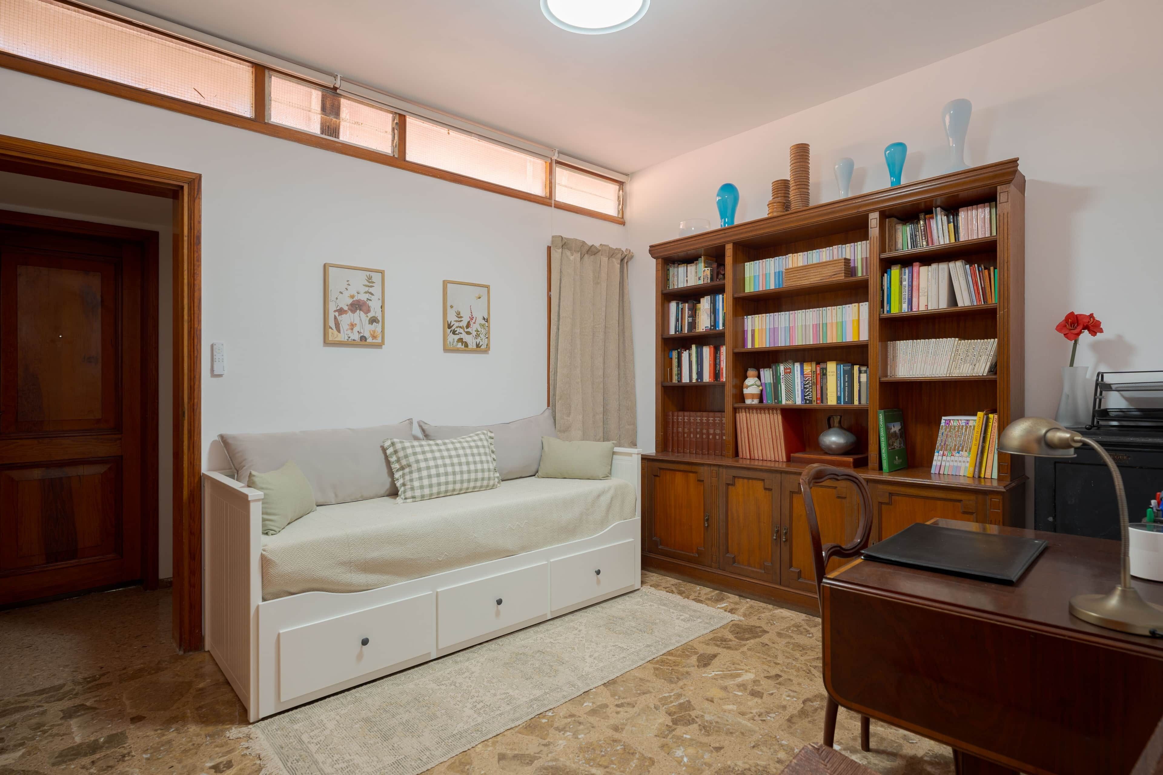 Una habitación con un sofá blanco, una estantería de madera, un escritorio con una lámpara y un suelo con diseño de piedra.