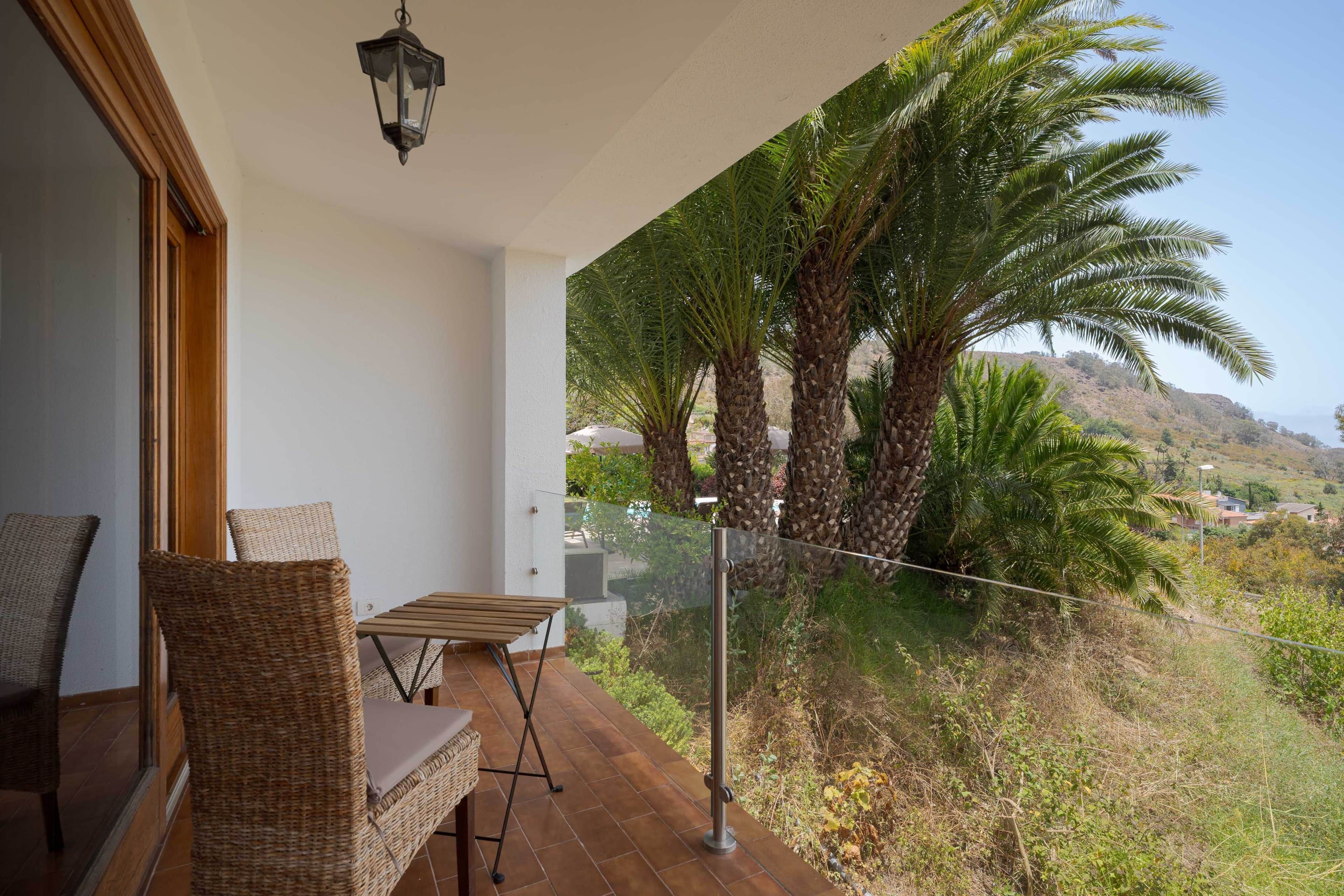 A balcony with wicker furniture, a table, and a view of palm trees and a hilly landscape.