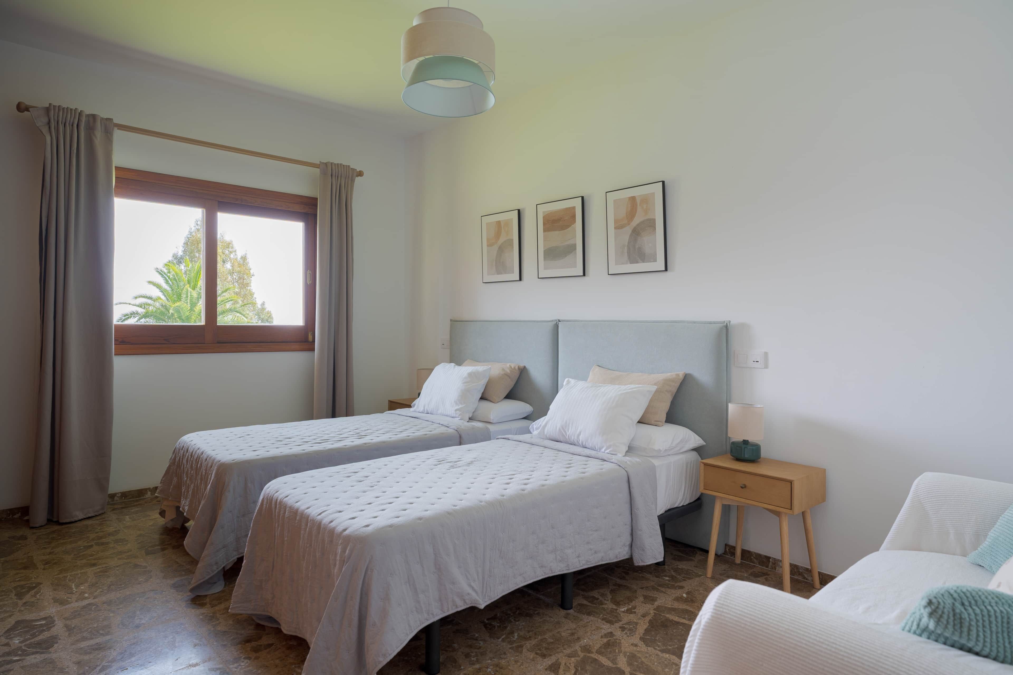 Une chambre avec deux lits, une table de chevet, une fenêtre avec des rideaux et des tableaux encadrés au mur.