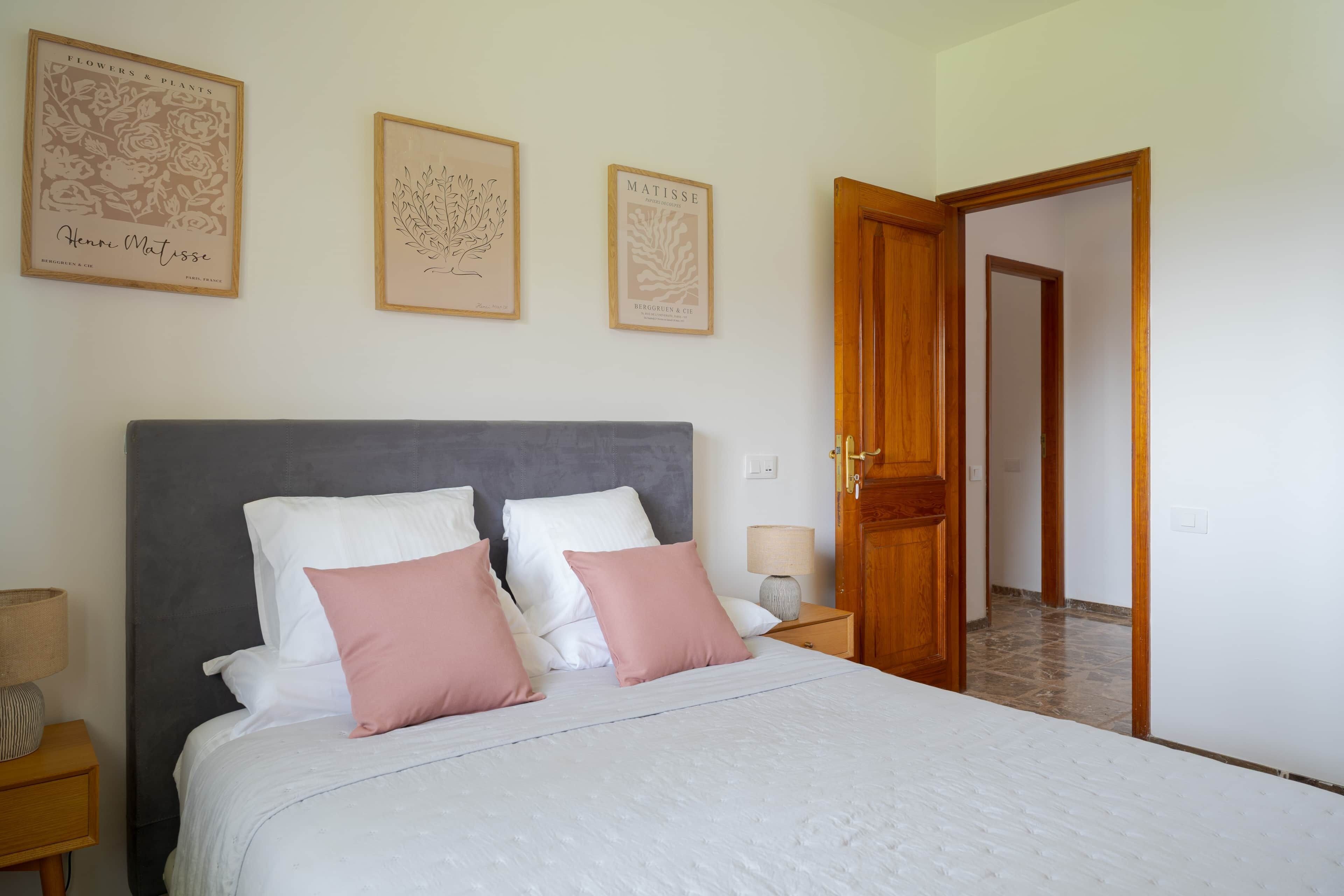 A bedroom with a bed, pillows, a nightstand, a lamp, and framed artwork on the wall.