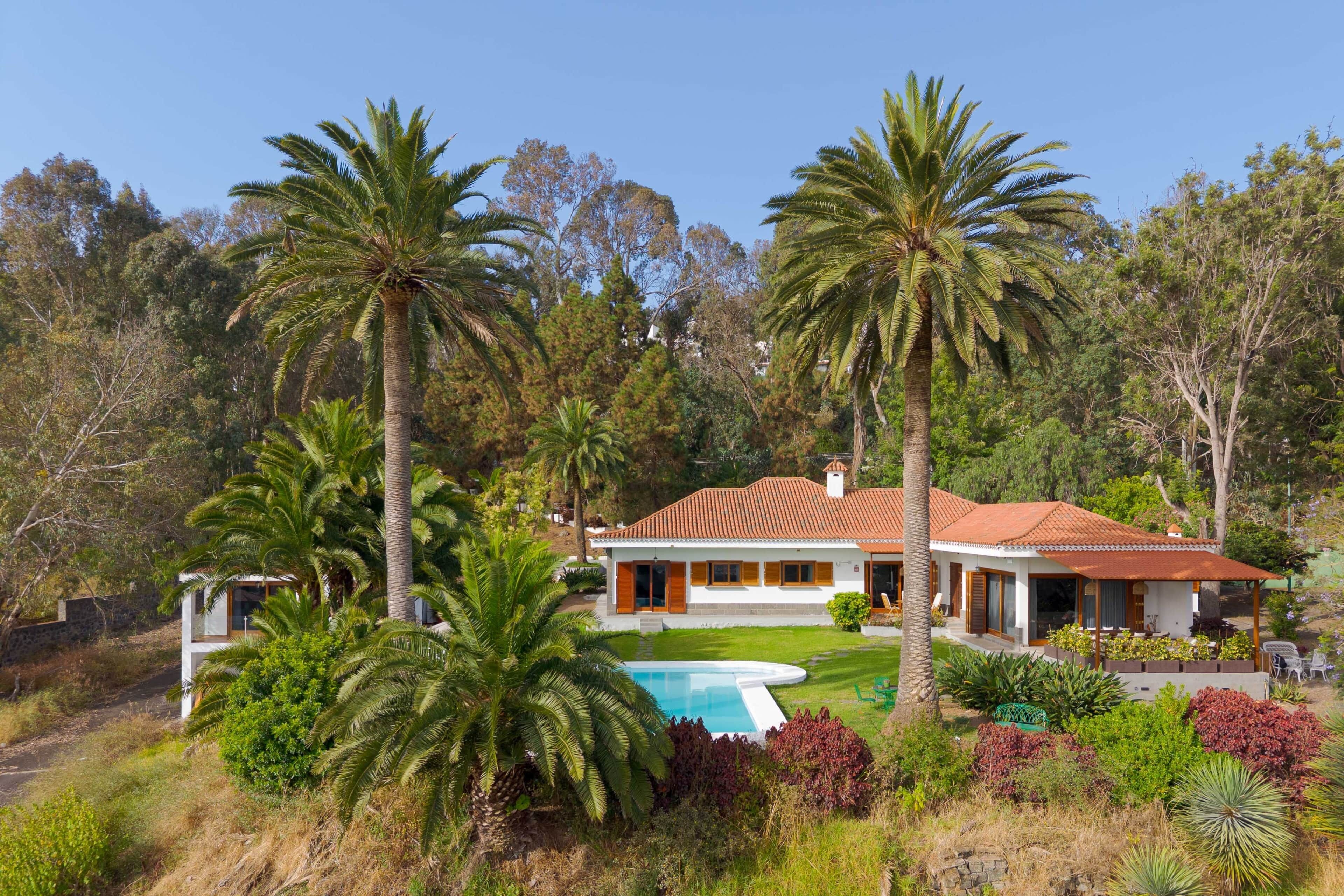 A two-story house with a swimming pool, surrounded by palm trees and a variety of vegetation.