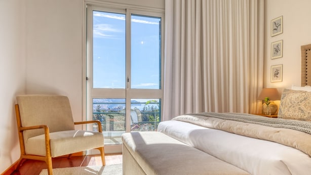 A bedroom with a large window, a chair, a bed, and a nightstand with a lamp. - Funchal Infinity, a Home in Madeira (Funchal)